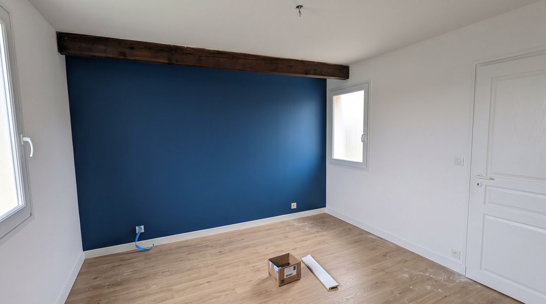 Chambre après travaux : mur peint en bleu profond, poutre bois conservée et remise en teinte, sol stratifié clair et plinthes blanches