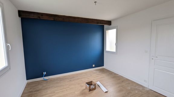 Chambre après travaux : mur peint en bleu profond, poutre bois conservée et remise en teinte, sol stratifié clair et plinthes blanches
