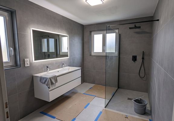 Salle de bain terminée : carrelage gris grand format au mur et au sol, douche à l'italienne avec paroi vitrée, meuble suspendu double vasque et miroir rétroéclairé
