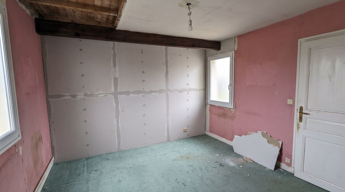 Chambre en cours de dépose : murs roses défraîchis, moquette verte, doublage en plaques de plâtre déjà posé sur un mur, poutre bois apparente