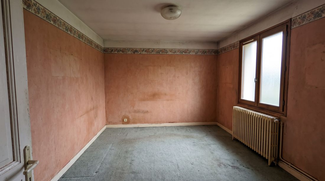 Chambre avant travaux : murs tapissés de papier peint saumon avec frise fleurie, moquette usée au sol, ancien radiateur en fonte
