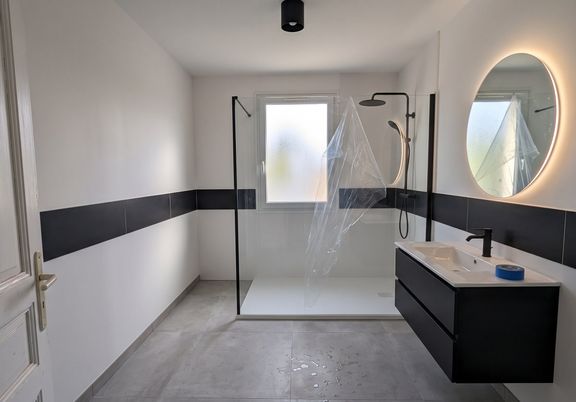 Salle de bain claire en fin de chantier : murs blancs avec bandeau de carrelage noir, receveur extra-plat et paroi de douche, meuble vasque noir et miroir rond rétroéclairé
