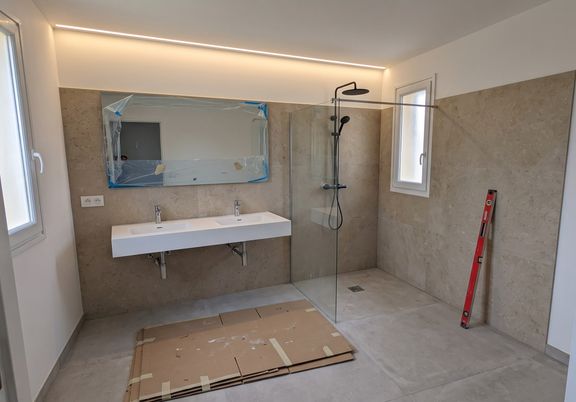Salle de bain en fin de chantier : carrelage effet pierre beige, plan double vasque suspendu, douche à l'italienne avec paroi vitrée et corniche lumineuse au plafond