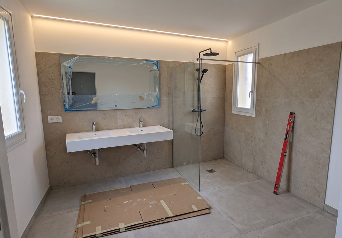 Salle de bain en fin de chantier : carrelage effet pierre beige, plan double vasque suspendu, douche à l'italienne avec paroi vitrée et corniche lumineuse au plafond
