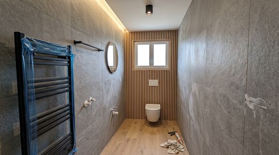 Petite salle d'eau : murs en carrelage effet pierre, habillage en tasseaux bois derrière un WC suspendu, sèche-serviettes et sol en lames bois
